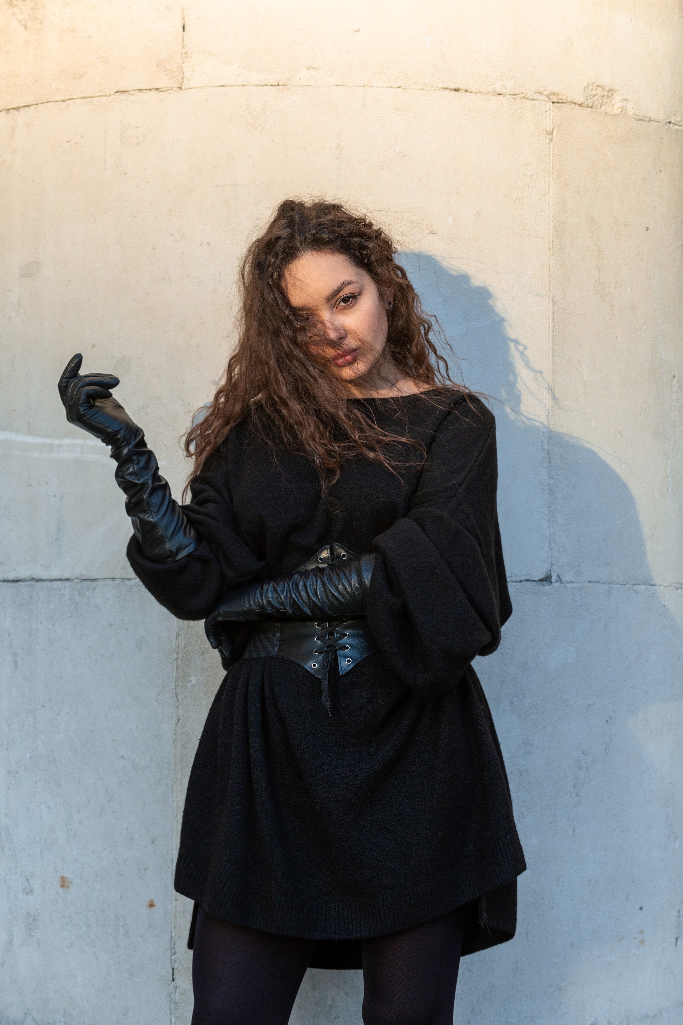Portret kobiety z kręconymi włosami w czarnej sukience, skórzanym gorsecie i długich rękawicach, pozującej na tle betonowej ściany.