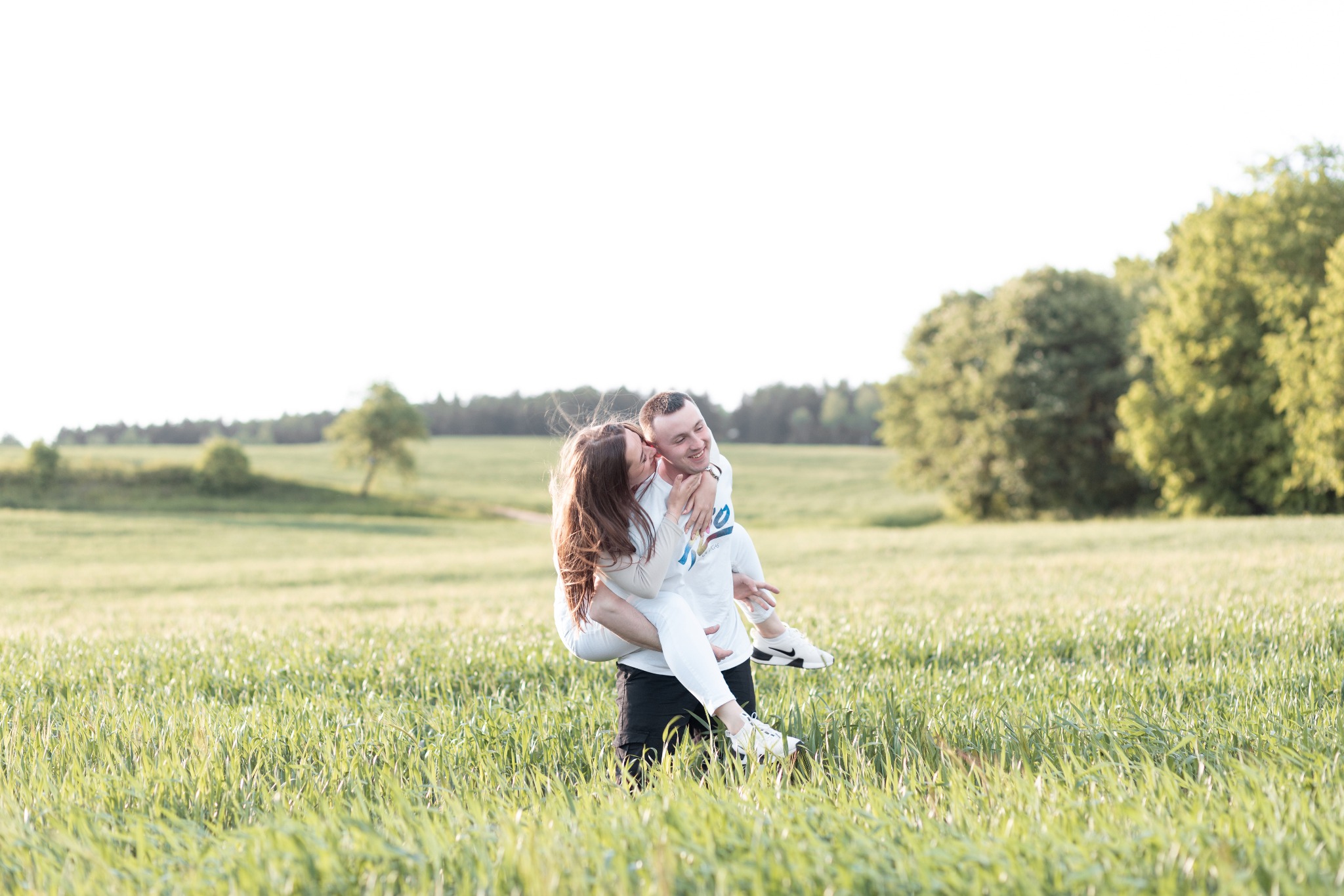 Uśmiechnięta para na łące: mężczyzna trzyma kobietę na barana, oboje ubrani na jasno, w tle zieleń drzew.