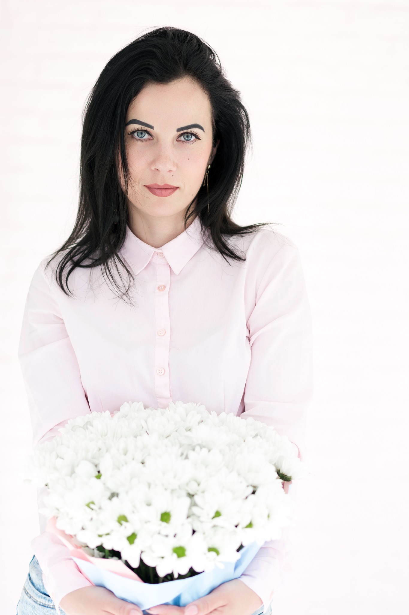 Portret kobiety o ciemnych włosach i niebieskich oczach, w jasnoróżowej koszuli, trzymającej bukiet białych chryzantem na jasnym tle.