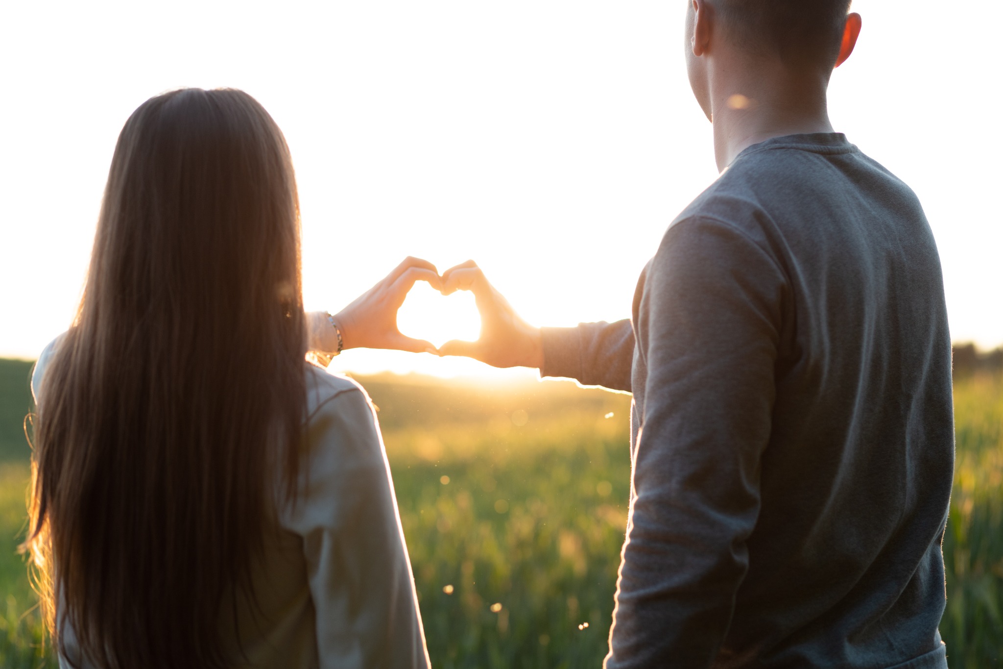 Para stojąca na tle zachodzącego słońca, z dłońmi ułożonymi w kształt serca, przez które przeświecają promienie słoneczne, widok od tyłu na łące.
