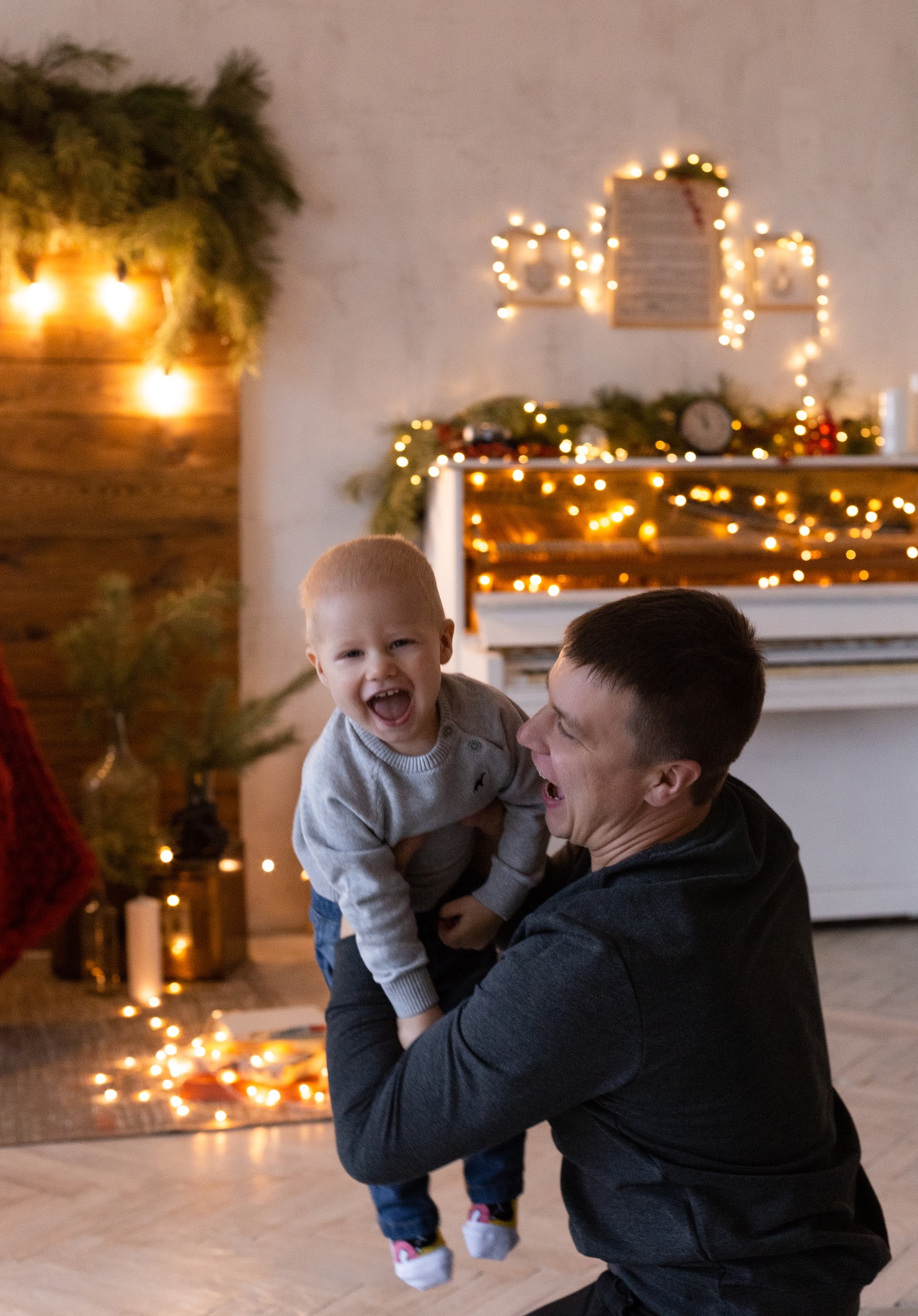 Uśmiechnięty ojciec trzymający na rękach roześmianego synka na tle świątecznego wystroju z pianinem, lampkami i zielonymi gałązkami.