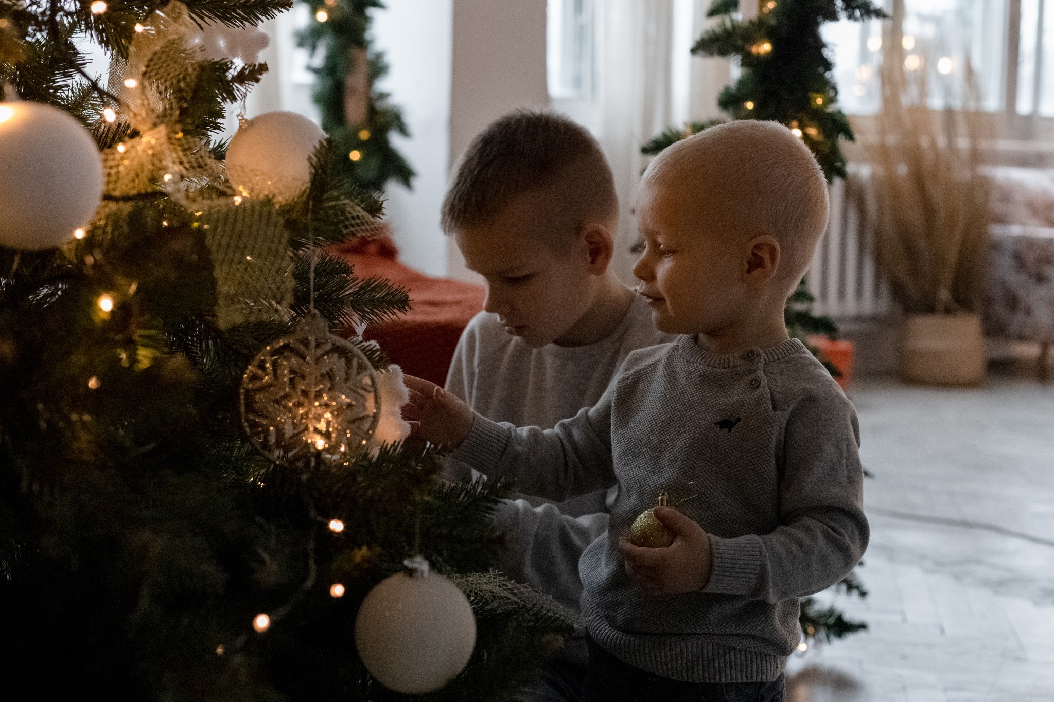 Dwóch chłopców w szarych swetrach dekoruje choinkę bombkami i światełkami w jasnym wnętrzu.