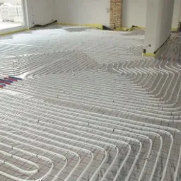 Rozłożone rury grzewcze na siatce w trakcie montażu ogrzewania podłogowego w jasnym pomieszczeniu z widocznymi elementami instalacji i żółtą taśmą zabezpieczającą wzdłuż ścian.