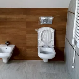 Nowa łazienka z drewnopodobną okładziną ścienną, wiszącą miską WC w folii ochronnej, bidetem i grzejnikiem drabinkowym.