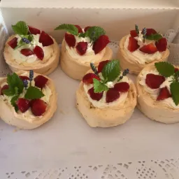 Sześć mini bez pavlova w kartonowym pudełku, udekorowanych bitą śmietaną, świeżymi truskawkami, listkami mięty i gałązką lawendy.