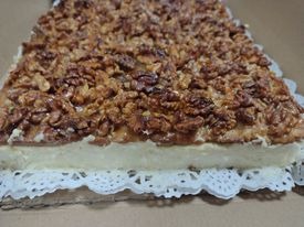 Kwadratowy tort orzechowy z kremową warstwą, udekorowany obficie połamanymi orzechami włoskimi, na białej, ażurowej podkładce.