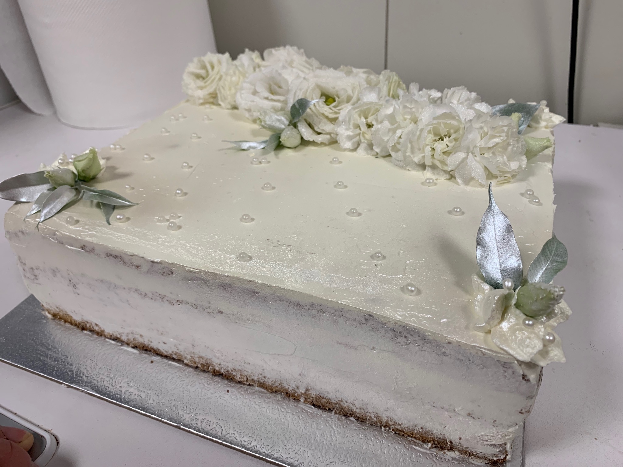 Prostokątny tort z białym kremem, dekorowany rzędem białych kwiatów i srebrnymi listkami oraz drobnymi perłami.