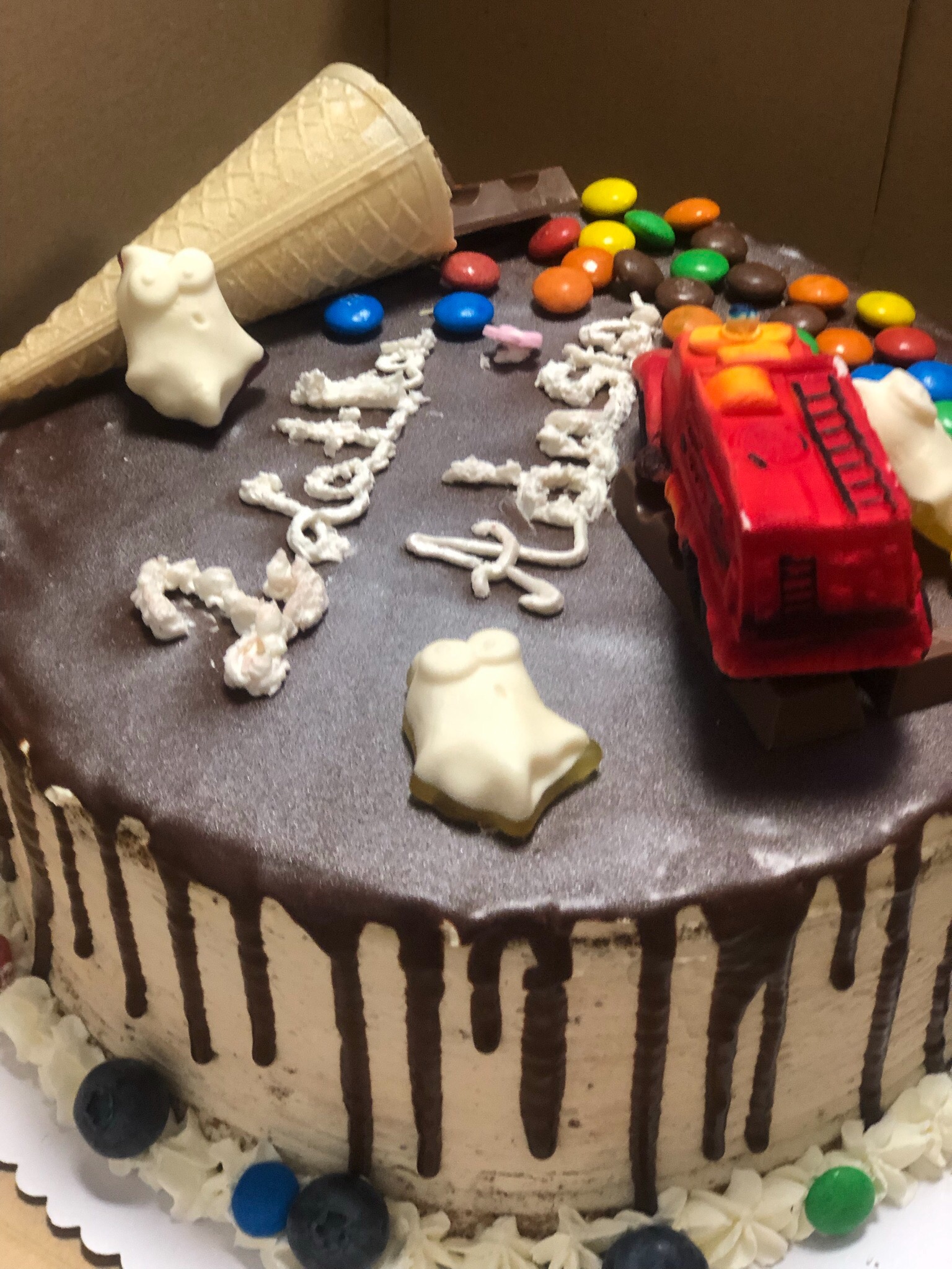Tort urodzinowy z czekoladową polewą, dekorowany drażami, wafelkiem, figurkami i zabawką w kształcie wozu strażackiego.