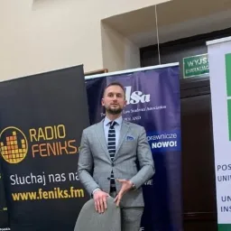 Mężczyzna w szarym garniturze i krawacie w paski, stojący na tle roll-upów reklamowych, w tym Radio Feniks i Koło Naukowe Postępowania Uniwersytetu Rzeszowskiego.