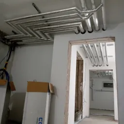 Instalacja wentylacyjna z rur owiniętych folią aluminiową, ułożonych zygzakiem pod sufitem w surowym wnętrzu, widoczne kartony z napisem 'CONEL CONNECTING ELEMENTS'.