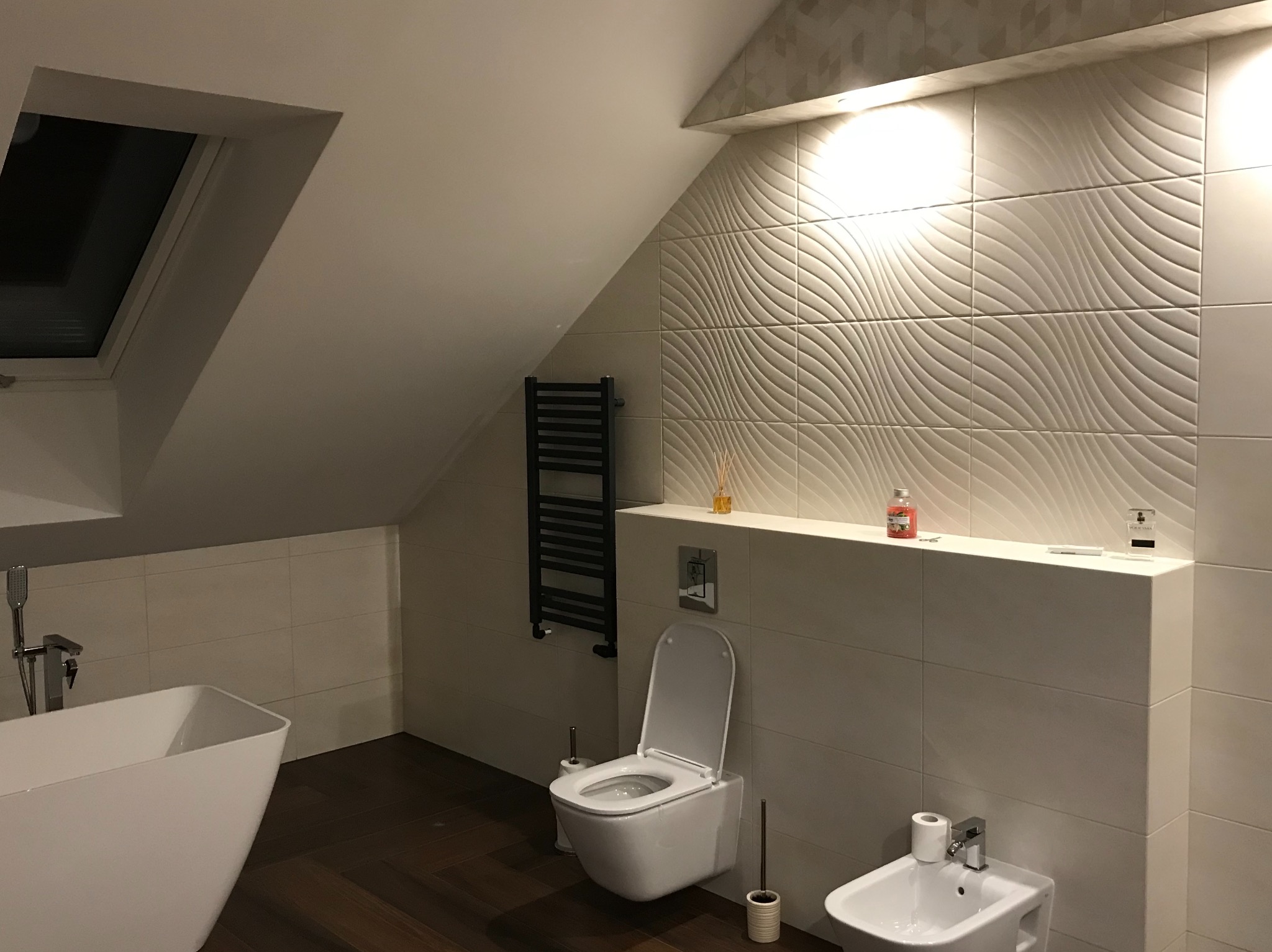 Nowoczesna łazienka na poddaszu z białą armaturą, dekoracyjnymi płytkami ściennymi z motywem fal, ciemną drewnianą podłogą i grzejnikiem drabinkowym w kolorze czarnym.