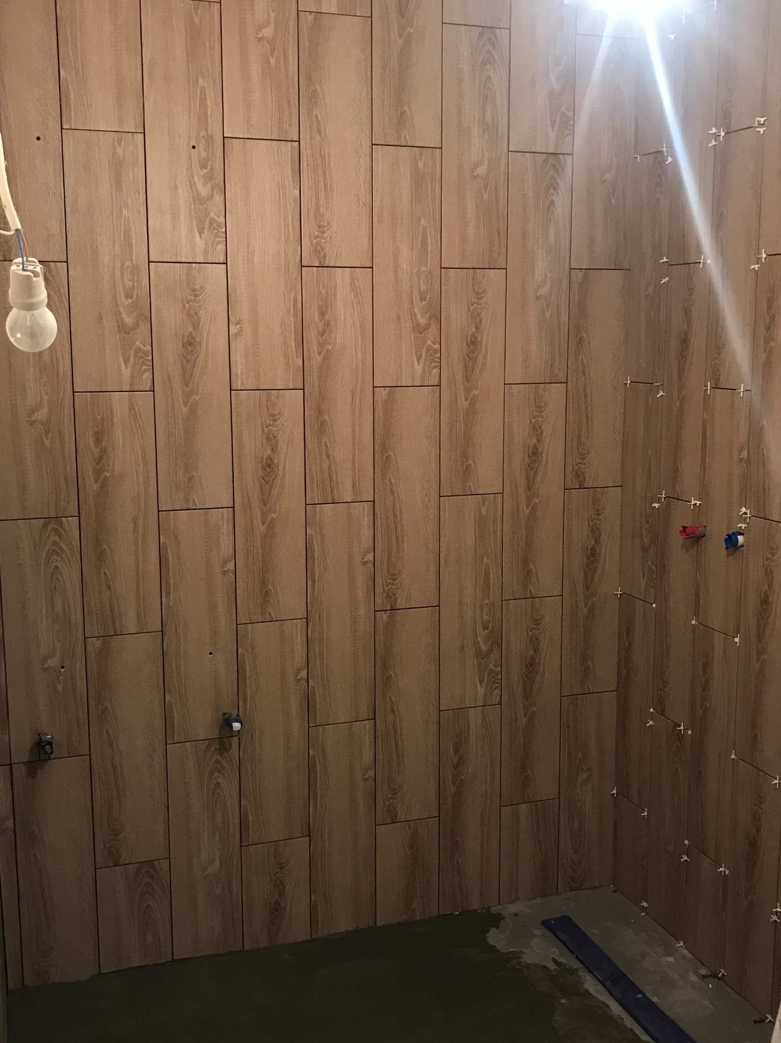 Ściana łazienki w trakcie układania płytek imitujących drewno, widoczne krzyżyki dystansowe, wystające kable z żarówką i fragment instalacji wodnej