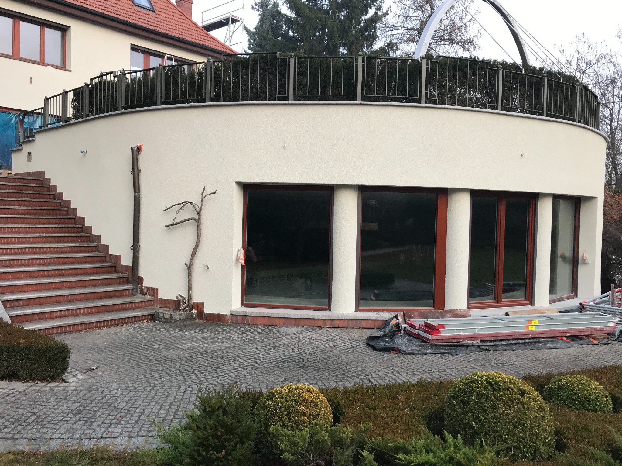 Elewacja domu po remoncie z nową, jasną gładzią, widoczne schody z czerwonej cegły, okna z drewnianymi ramami i metalowe rusztowanie na posesji.