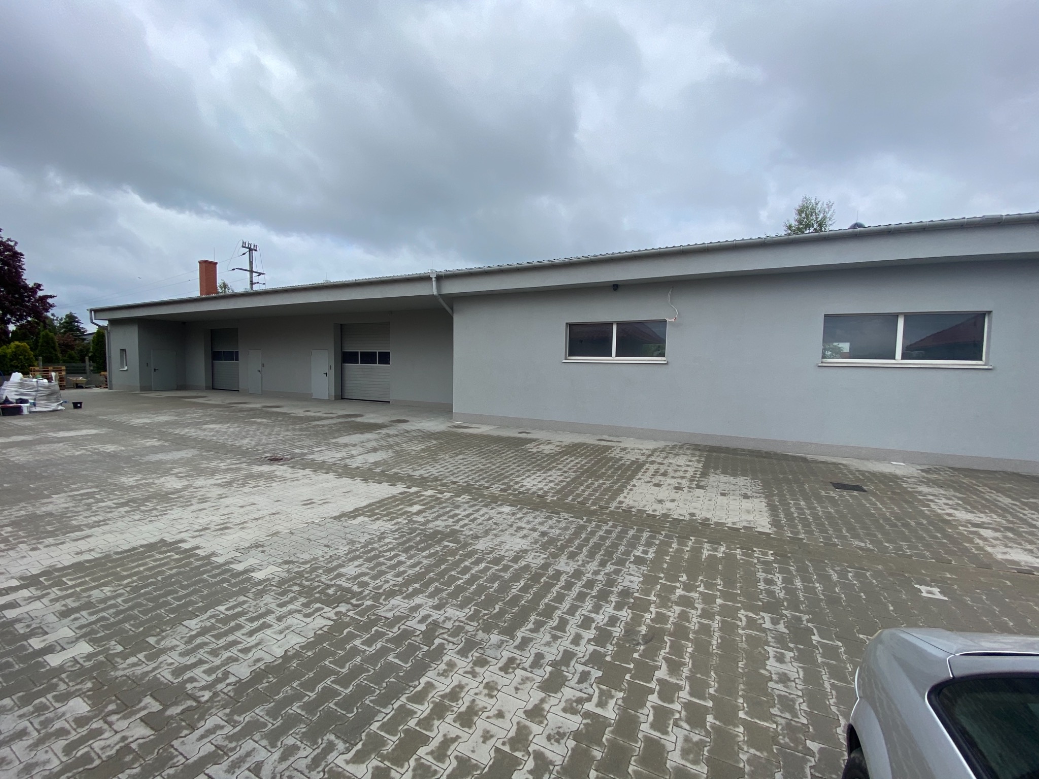 Szary budynek z bramami garażowymi i dwoma oknami, świeżo pomalowany, z mokrą kostką brukową przed budynkiem w pochmurny dzień.