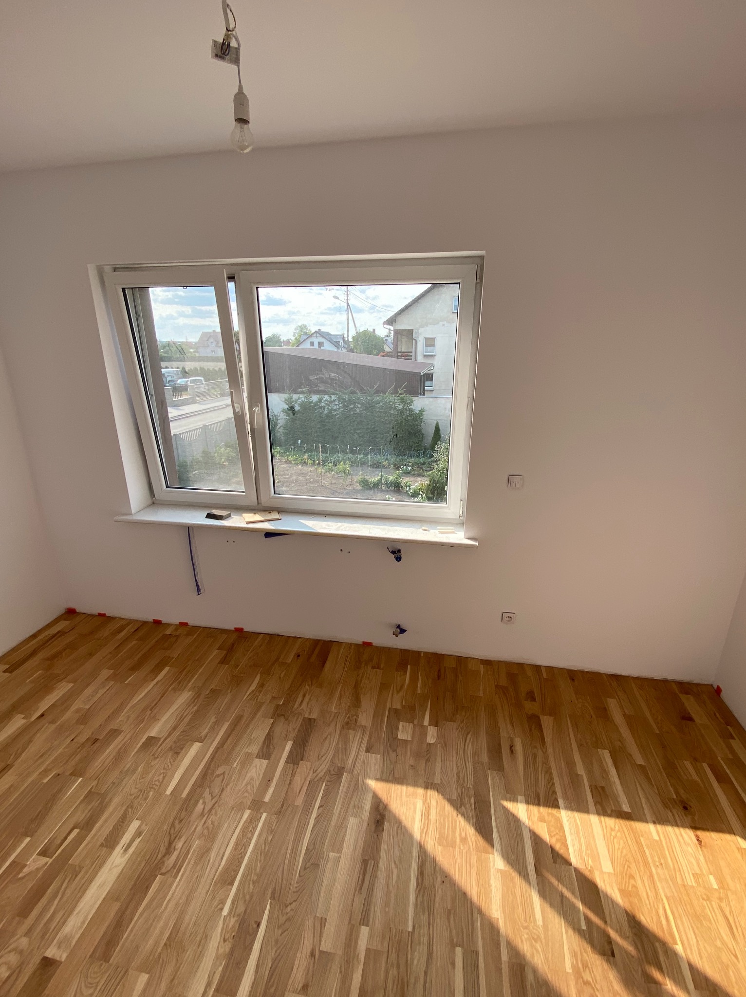 Jasny pokój po remoncie z nowo ułożonym parkietem w jodełkę, widoczne listwy przypodłogowe zabezpieczone taśmą, surowa żarówka zwisająca z sufitu i okno z widokiem na ogród.