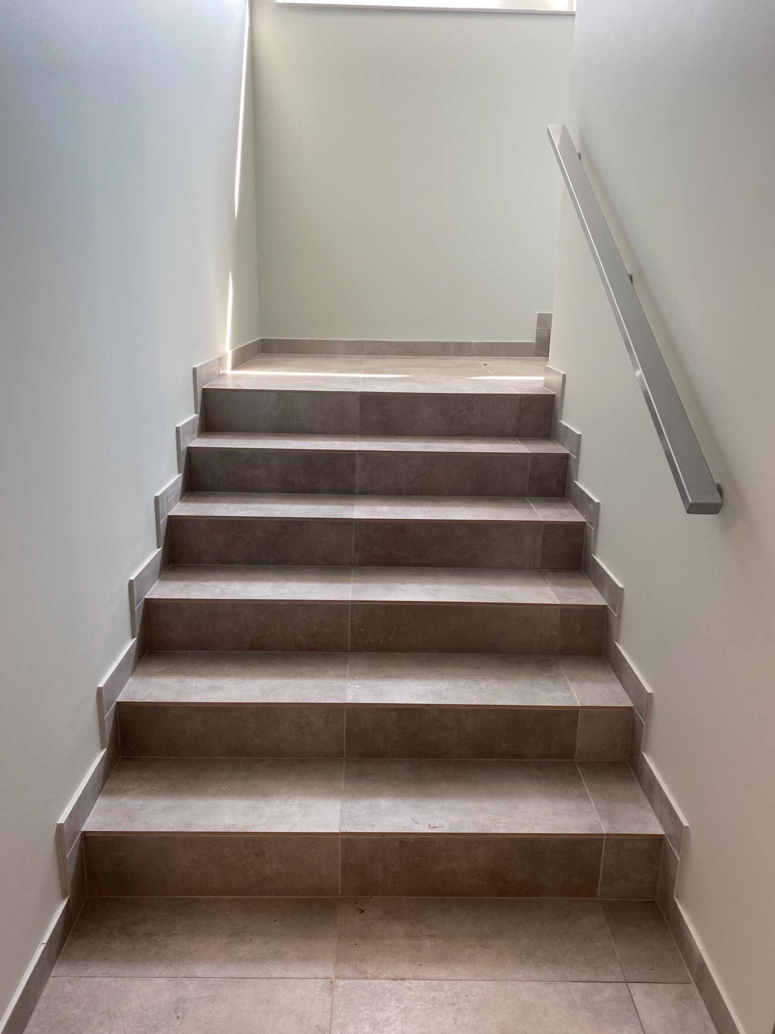 Schody wewnętrzne wyłożone brązowymi płytkami ceramicznymi, z metalową poręczą po prawej stronie i jasnym oświetleniem wpadającym z góry.