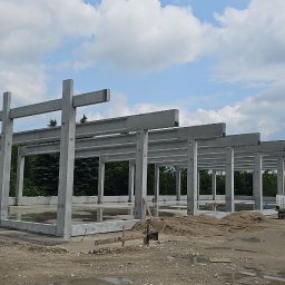 BAUPOL - NADZORY BUDOWLANE - Surowy szkielet budynku z betonu w trakcie budowy, widok z zewnątrz, z niebieskim podnośnikiem koszowym po lewej stronie. Niebo z chmurami w tle.