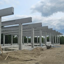 BAUPOL - NADZORY BUDOWLANE - Konstrukcja szkieletowa budynku z betonu, widok z zewnątrz, z placem budowy i zbiornikiem IBC w tle, pochmurne niebo.