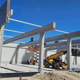 BAUPOL - NADZORY BUDOWLANE - Montaż paneli warstwowych na hali z wykorzystaniem podnośników. Widoczne betonowe słupy i pracownicy na wysokości. Słoneczny dzień, błękitne niebo.