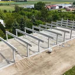 BAUPOL - NADZORY BUDOWLANE - Wysoka perspektywa na plac budowy z betonowymi słupami i belkami, przygotowany teren pod budowę, w tle zieleń i zabudowania. Widoczne ślady opon na ziemi.