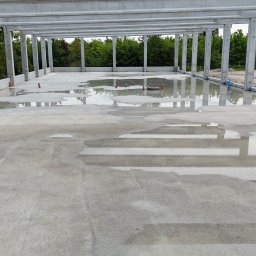 BAUPOL - NADZORY BUDOWLANE - Betonowa konstrukcja hali w stanie surowym otwartym, z widocznymi słupami i stropem, zalana wodą po deszczu, z instalacją sanitarną. Widok na zewnątrz.