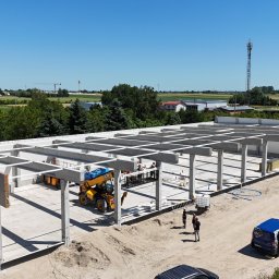 BAUPOL - NADZORY BUDOWLANE - Budowa hali z prefabrykatów betonowych w słoneczny dzień. Widoczny dźwig teleskopowy, pracownicy i elementy konstrukcyjne. W tle zieleń i zabudowania.