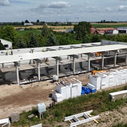 BAUPOL - NADZORY BUDOWLANE - Budowa hali z dachem i stalową konstrukcją w słoneczny dzień. Widoczni pracownicy na dachu, materiały budowlane i maszyny. W tle krajobraz z polami i budynkami.