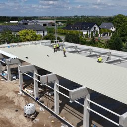 BAUPOL - NADZORY BUDOWLANE - Montaż dachu hali z betonu i stali. Widoczni pracownicy w żółtych kamizelkach, na tle zabudowań i zieleni. Ujęcie z góry ukazuje postęp prac.