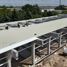 Montaż blachy trapezowej na dachu hali przemysłowej. Czterech robotnik&oacute;w w jaskrawych kamizelkach pracuje na dachu. Widok z g&oacute;ry na konstrukcję i okoliczny krajobraz.