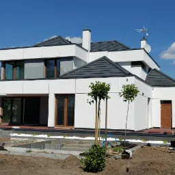 Nowoczesny, biały dom z szarą dachówką i minimalistycznym ogrodem w trakcie aranżacji, widoczne drzewka i ścieżka z kostki brukowej.