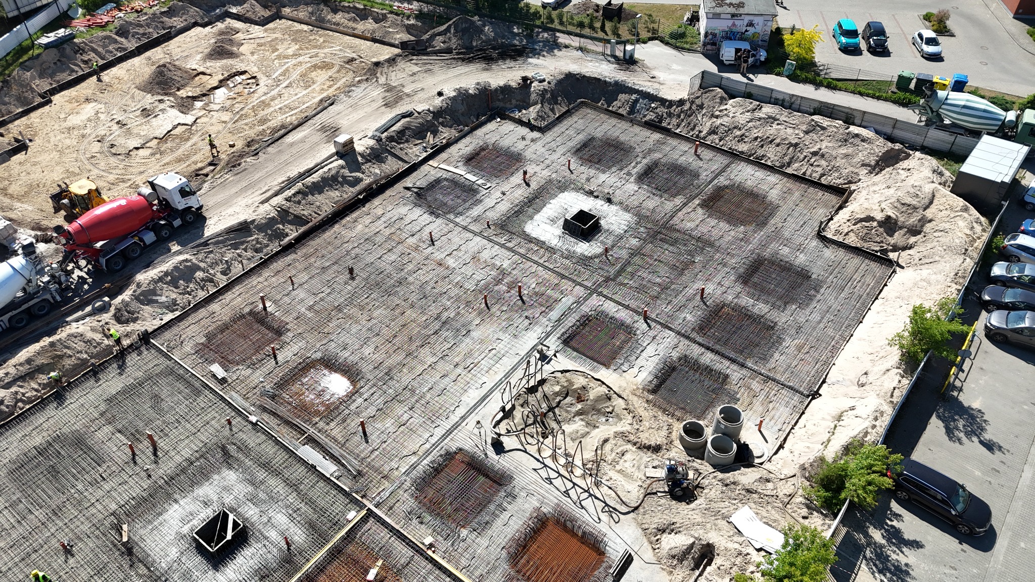 Widok z góry na plac budowy z przygotowaną zbrojoną płytą fundamentową, betonowe miksokrety i koparka w tle, teren częściowo wyrównany piaskiem.