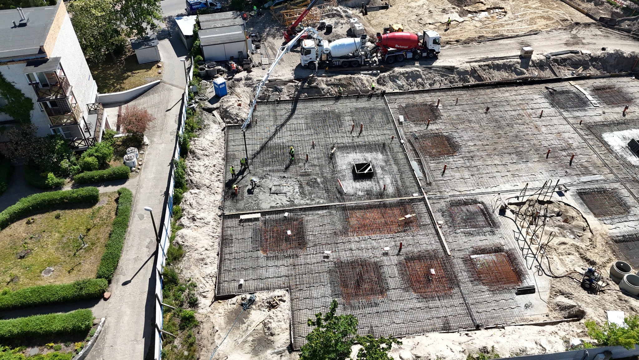Zalewanie betonem zbrojonej płyty fundamentowej budynku z użyciem pompy do betonu, widok z góry na plac budowy z fragmentem zieleni i zabudowań obok.