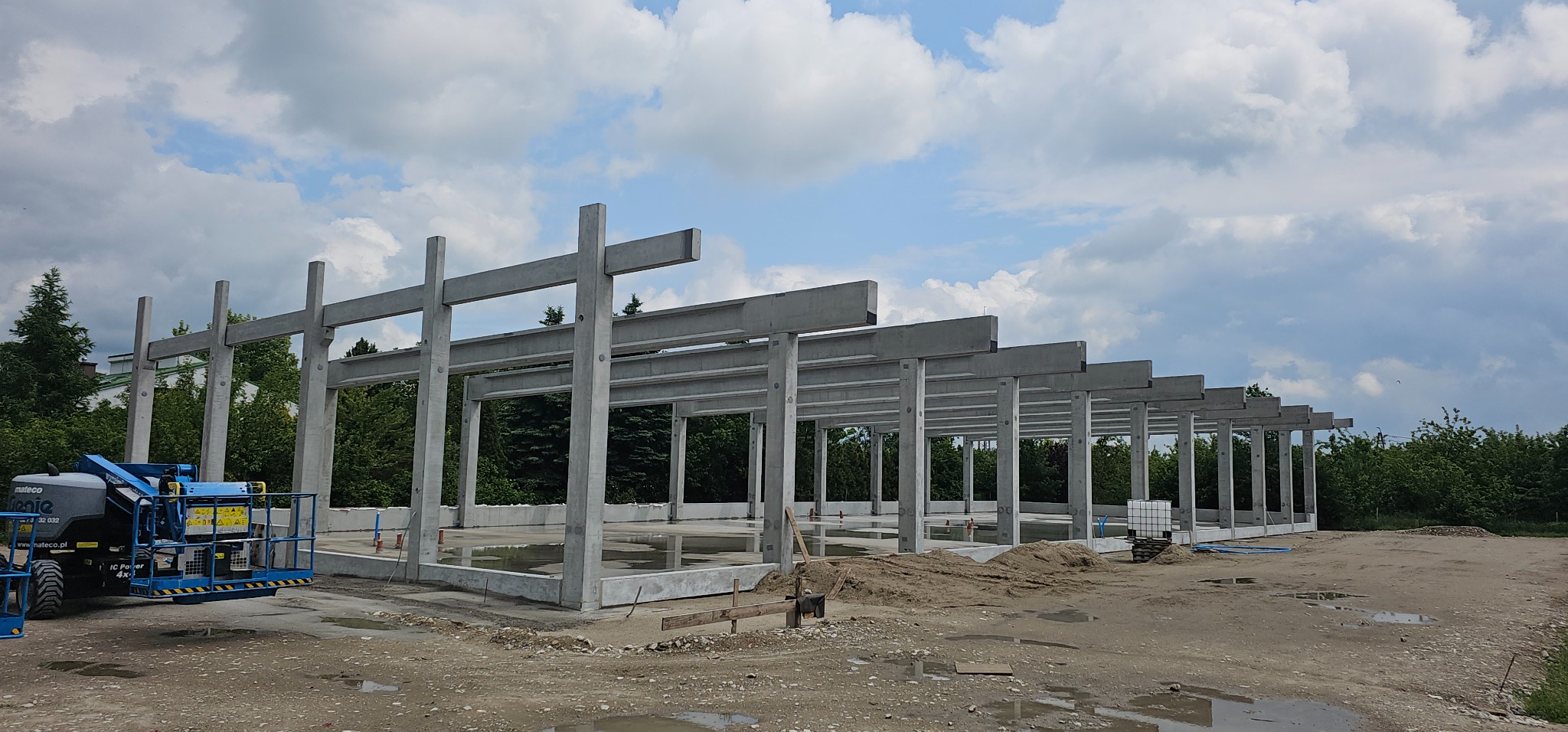 Surowy szkielet budynku z betonu w trakcie budowy, widok z zewnątrz, z niebieskim podnośnikiem koszowym po lewej stronie. Niebo z chmurami w tle.