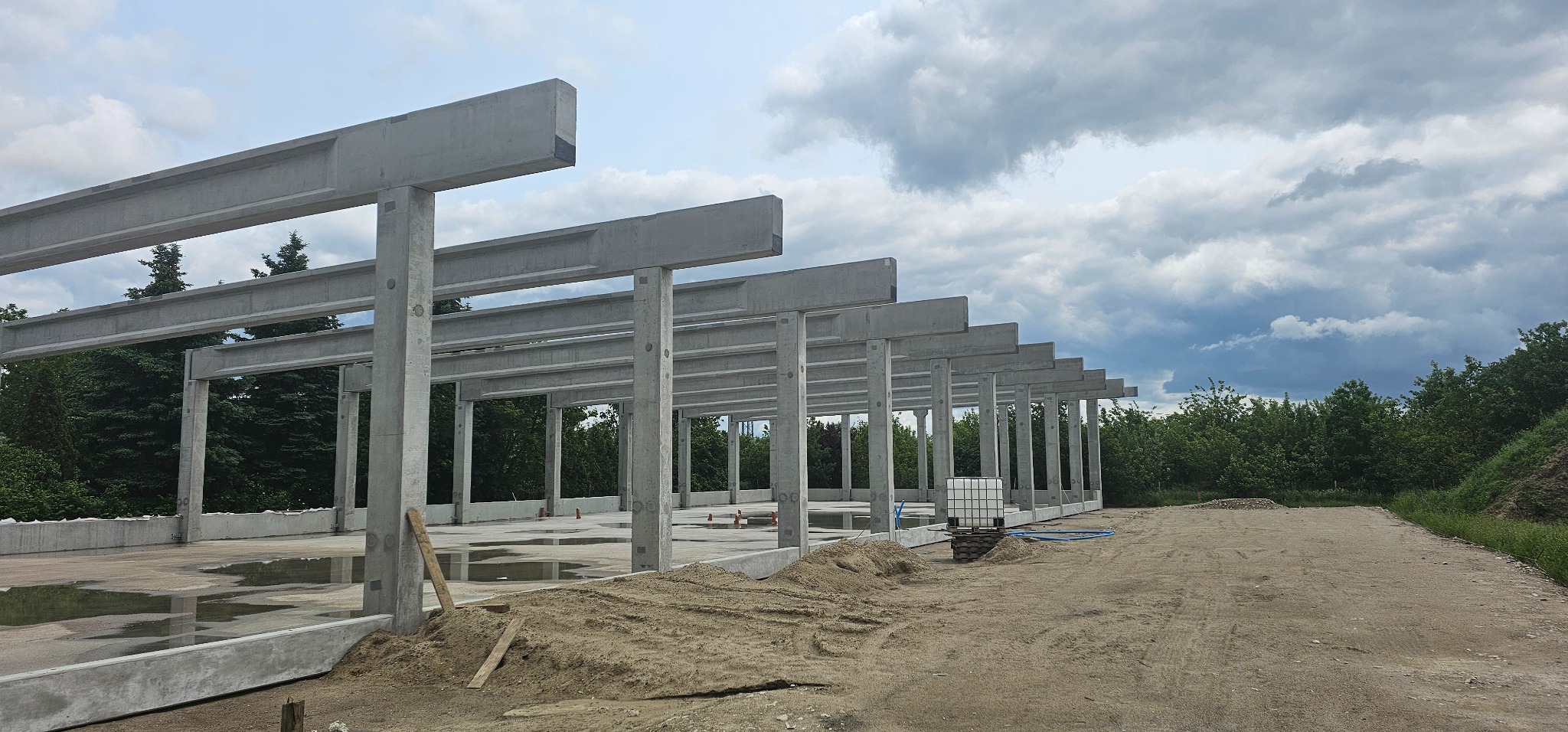 Konstrukcja szkieletowa budynku z betonu, widok z zewnątrz, z placem budowy i zbiornikiem IBC w tle, pochmurne niebo.