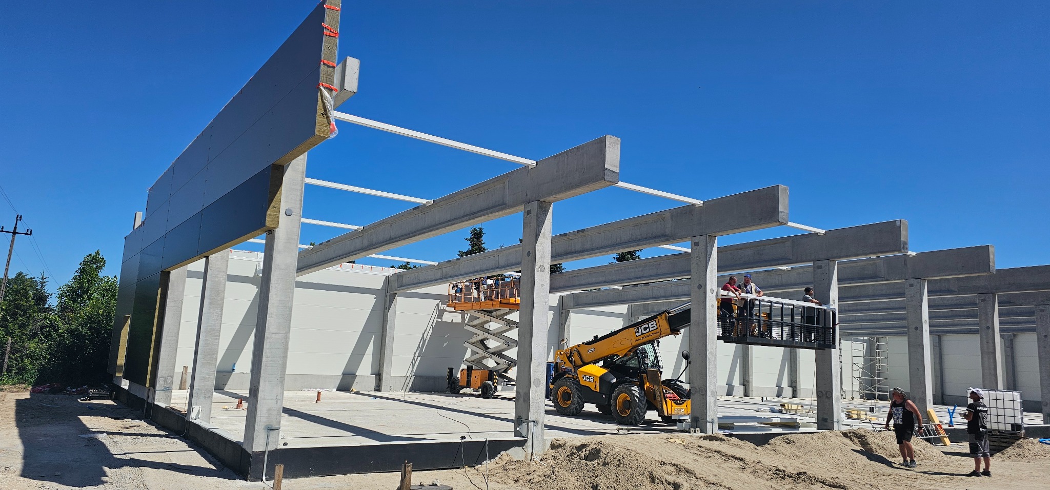 Montaż paneli warstwowych na hali z wykorzystaniem podnośników. Widoczne betonowe słupy i pracownicy na wysokości. Słoneczny dzień, błękitne niebo.