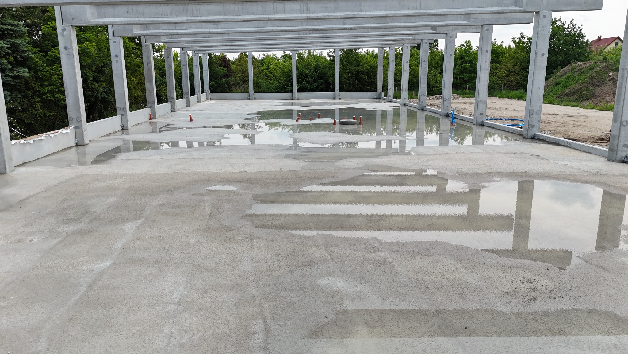 Betonowa konstrukcja hali w stanie surowym otwartym, z widocznymi słupami i stropem, zalana wodą po deszczu, z instalacją sanitarną. Widok na zewnątrz.