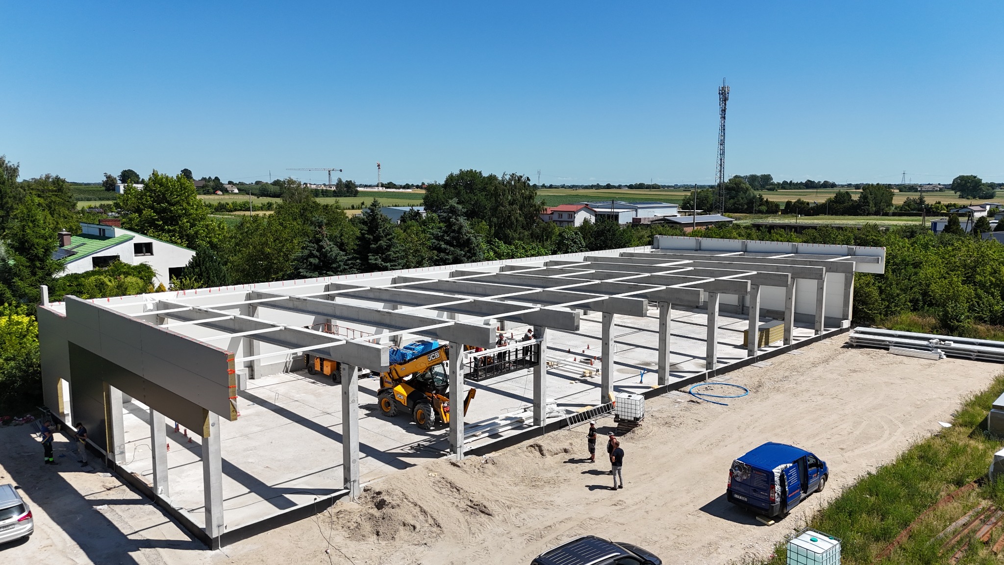 Budowa hali z prefabrykatów betonowych w słoneczny dzień. Widoczny dźwig teleskopowy, pracownicy i elementy konstrukcyjne. W tle zieleń i zabudowania.