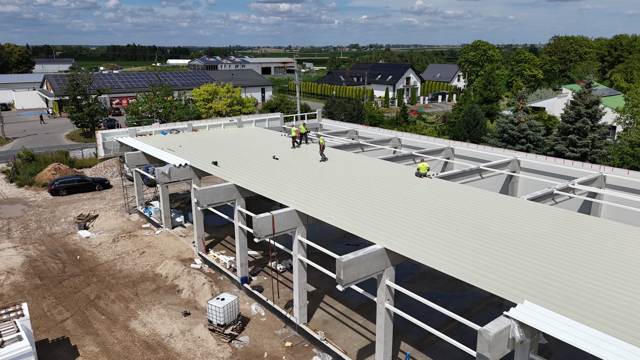 Montaż dachu hali z betonu i stali. Widoczni pracownicy w żółtych kamizelkach, na tle zabudowań i zieleni. Ujęcie z góry ukazuje postęp prac.