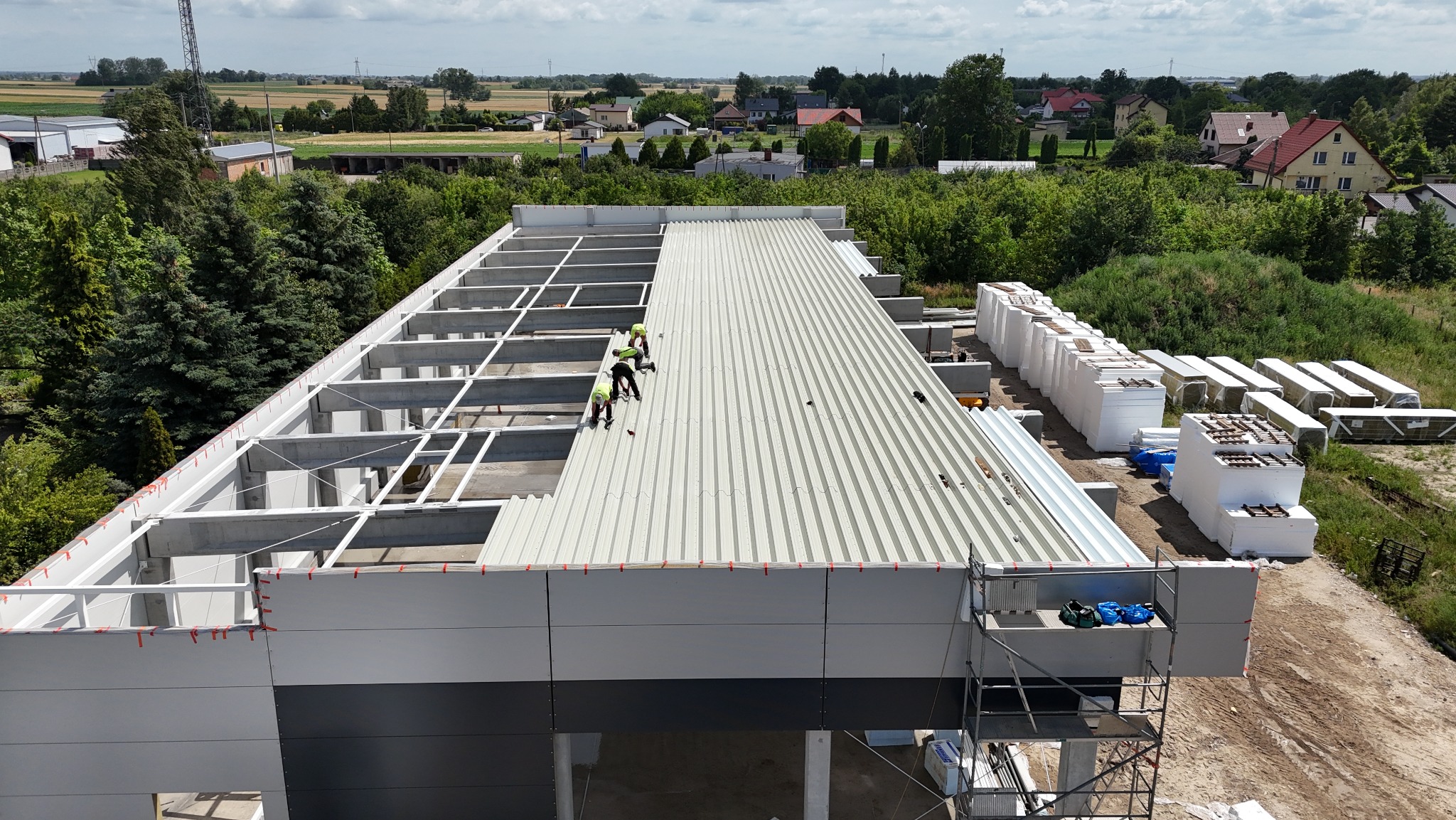 Montaż dachu z blachy trapezowej na hali przemysłowej. Prace na wysokości, widoczne materiały izolacyjne i konstrukcja szkieletowa. Ujęcie z góry na budowę w toku.