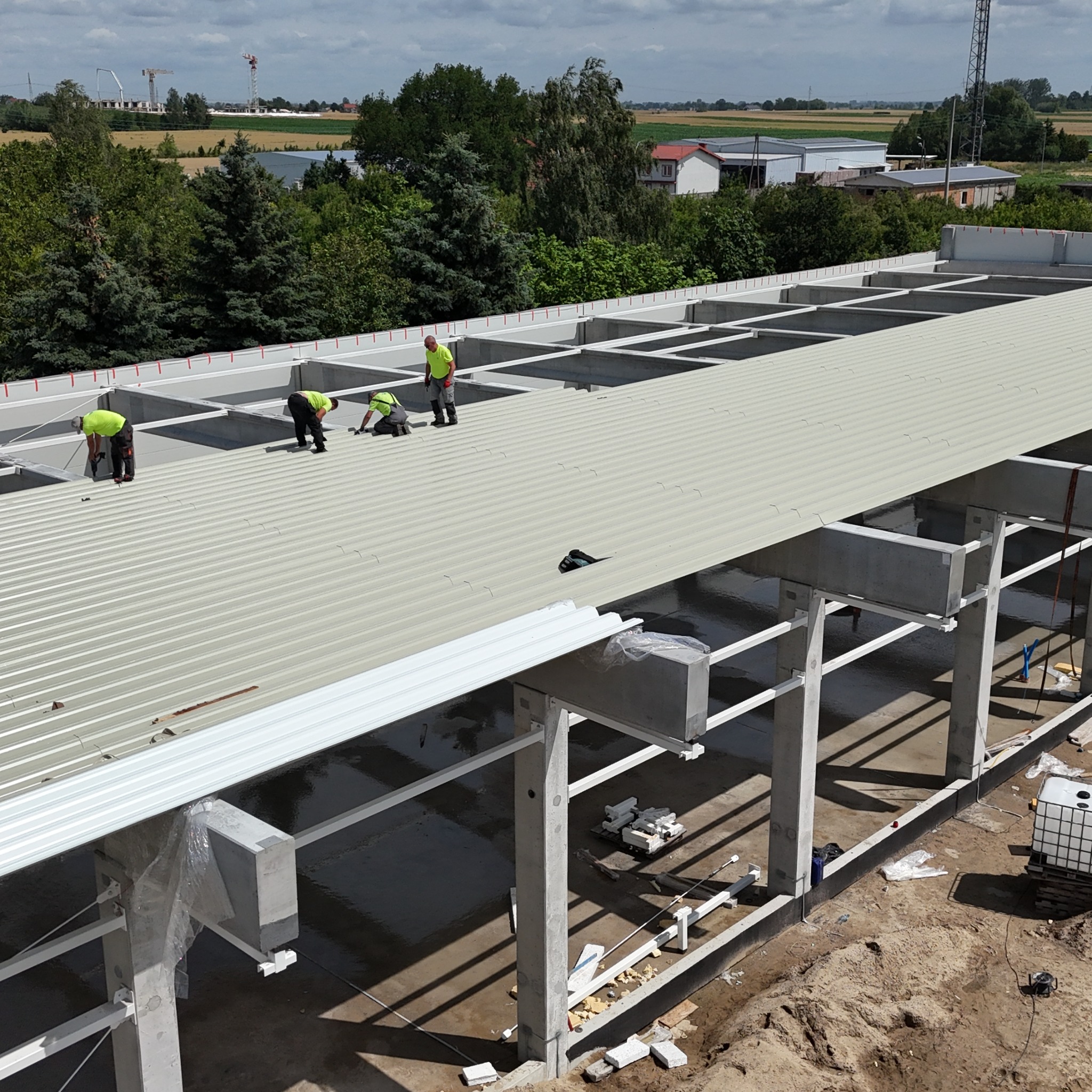 Montaż blachy trapezowej na dachu hali przemysłowej. Czterech robotników w jaskrawych kamizelkach pracuje na dachu. Widok z góry na konstrukcję i okoliczny krajobraz.