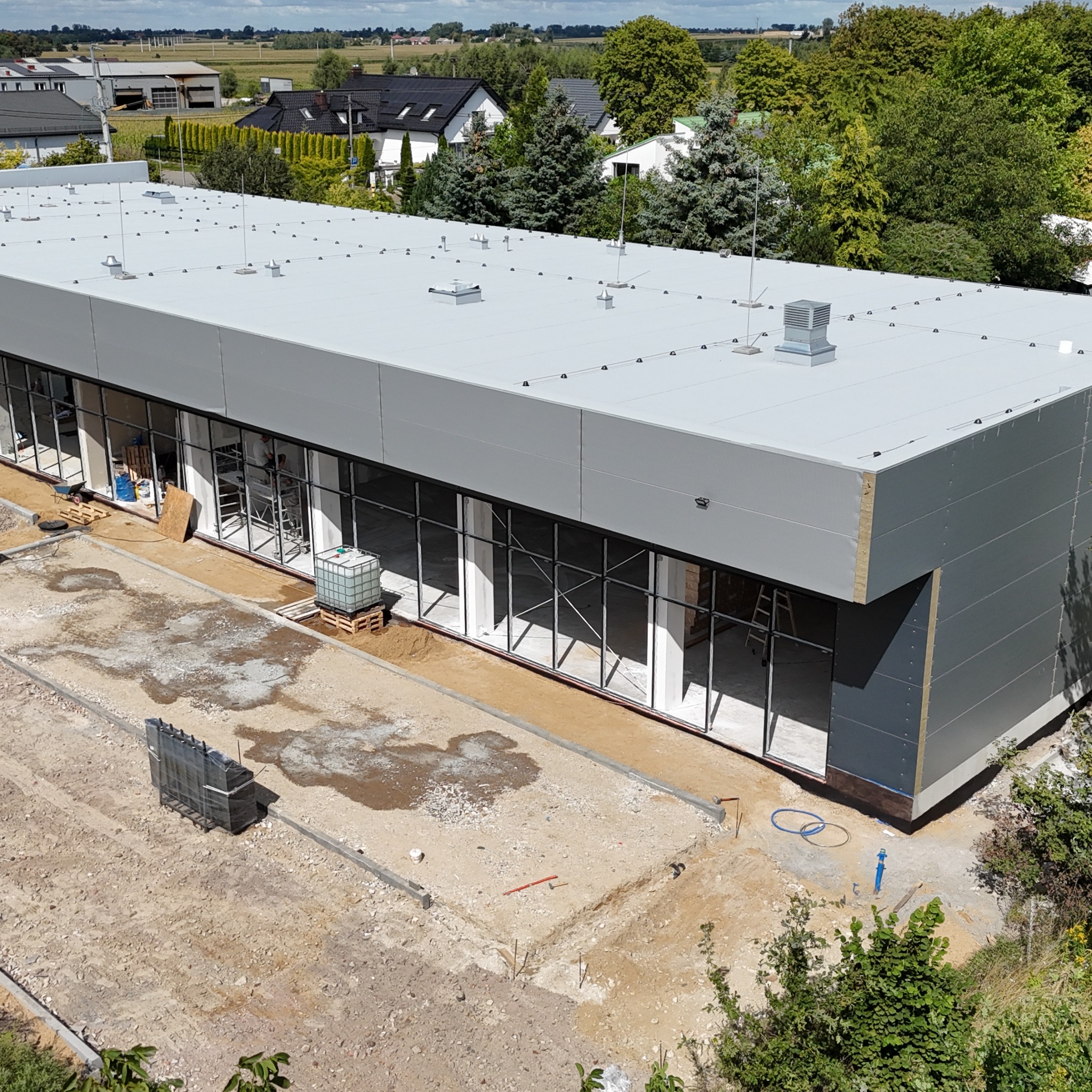 Budynek komercyjny w trakcie budowy: szara elewacja z dużymi oknami, widoczne rusztowania i materiały budowlane na zewnątrz. Ujęcie z góry.