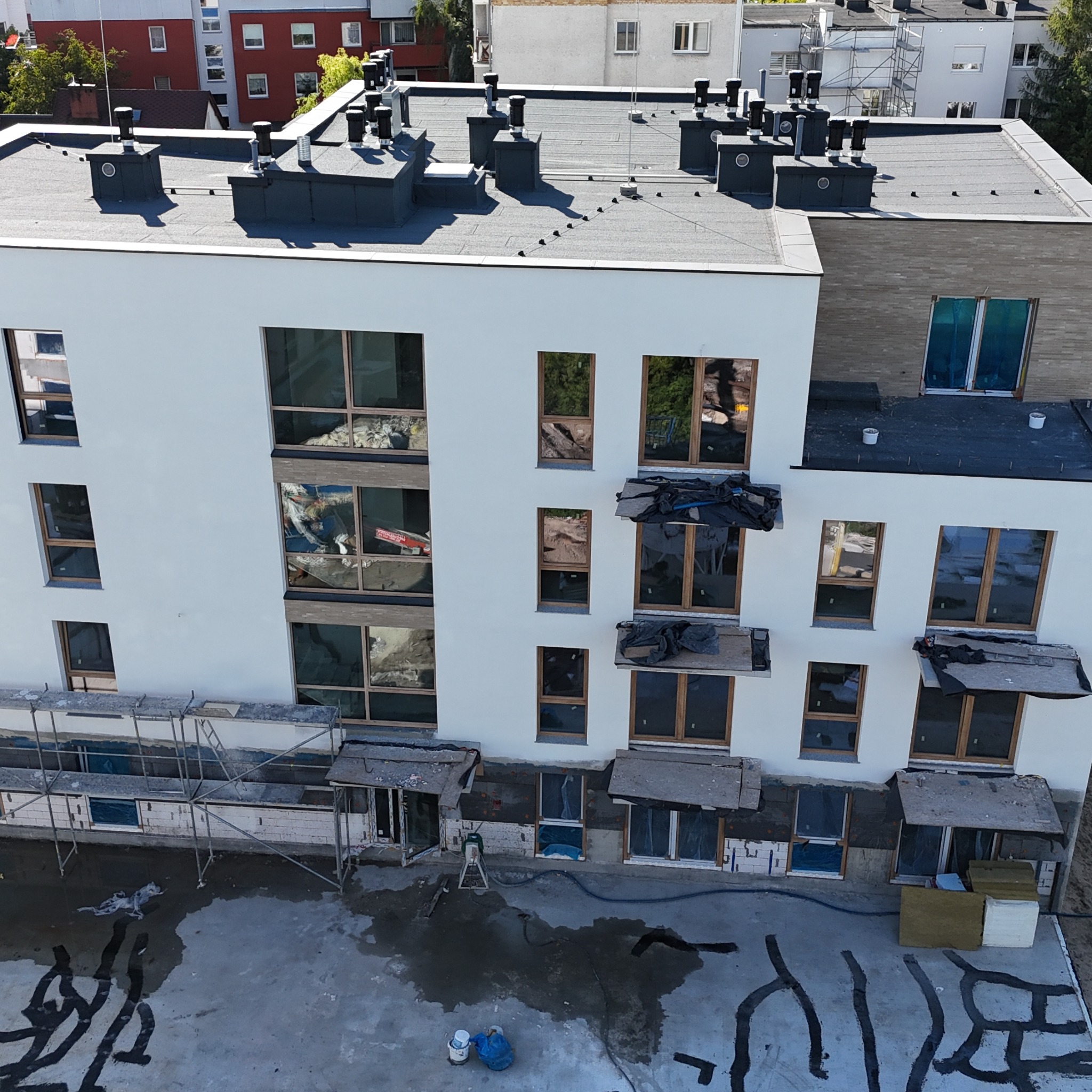 Wysoki kąt na budynek w trakcie ocieplania. Widoczne rusztowania, okna z odbiciami, izolacja i elementy wykończeniowe. Dach płaski z kominami.