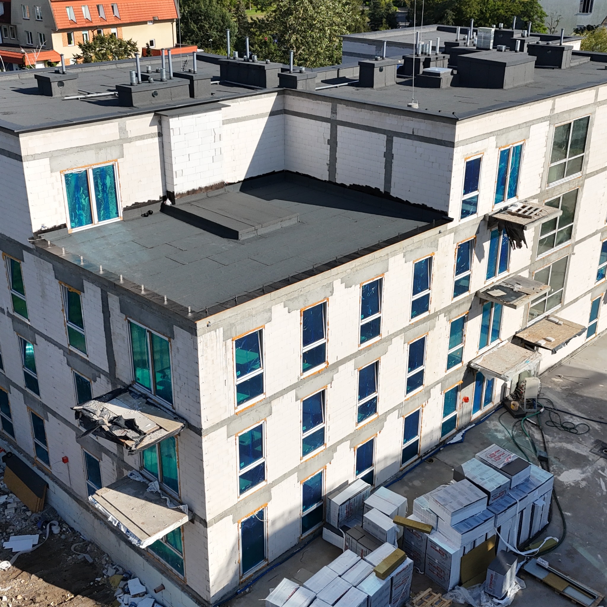 Wielopiętrowy budynek w stanie surowym zamkniętym z niebieską folią na oknach, widok z góry. Na dachu kominy, na placu budowy materiały budowlane.