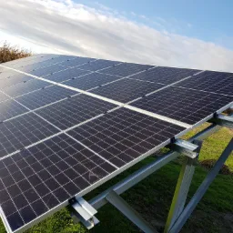 Panel słoneczny na metalowej konstrukcji, widok z dołu pod kątem, na tle zielonej trawy i błękitnego nieba z chmurami.