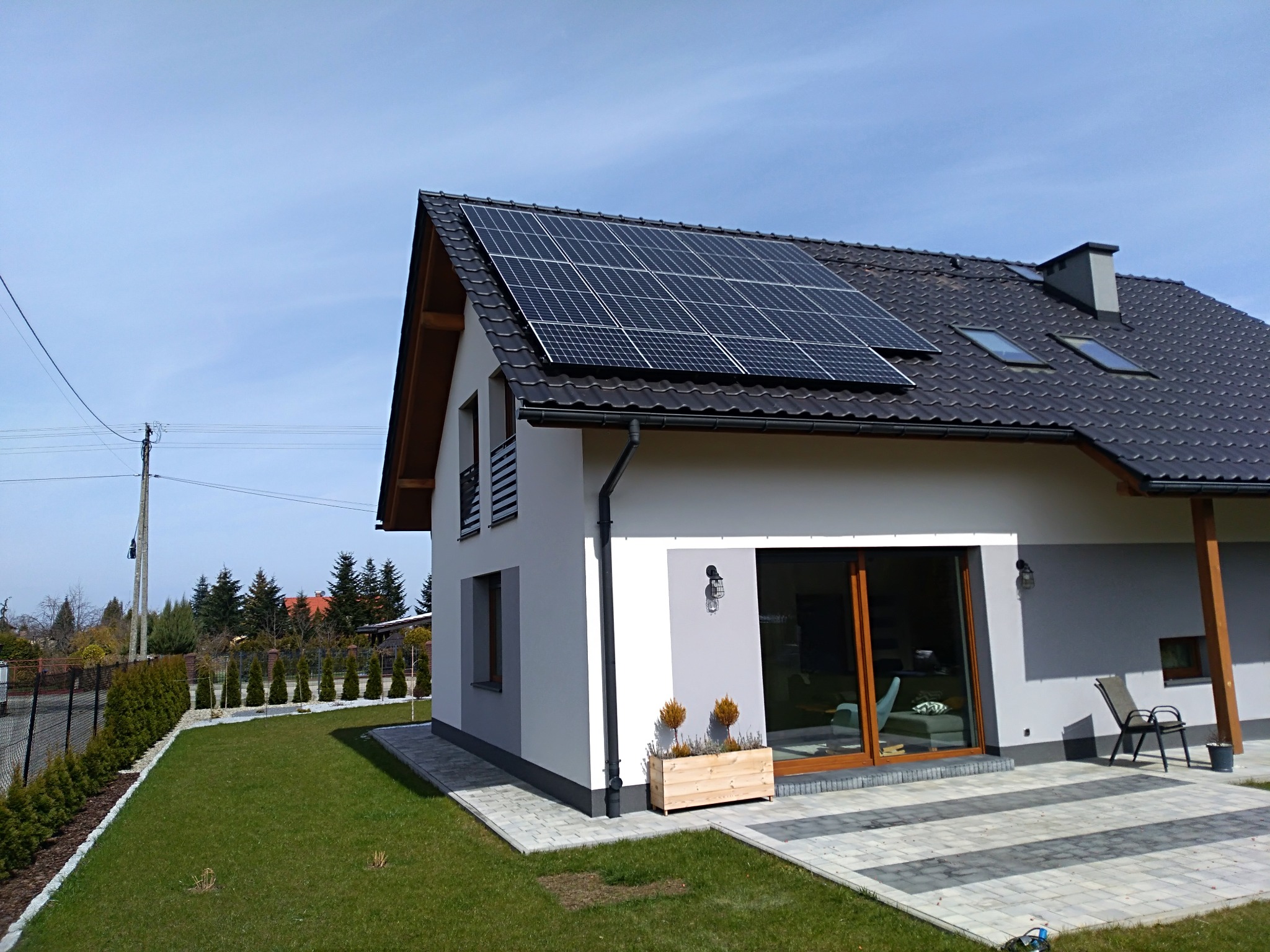 Nowoczesny dom jednorodzinny z panelami słonecznymi na ciemnej dachówce, widoczne okna dachowe i komin, zadbany trawnik, tuje wzdłuż ogrodzenia, jasne elewacje.