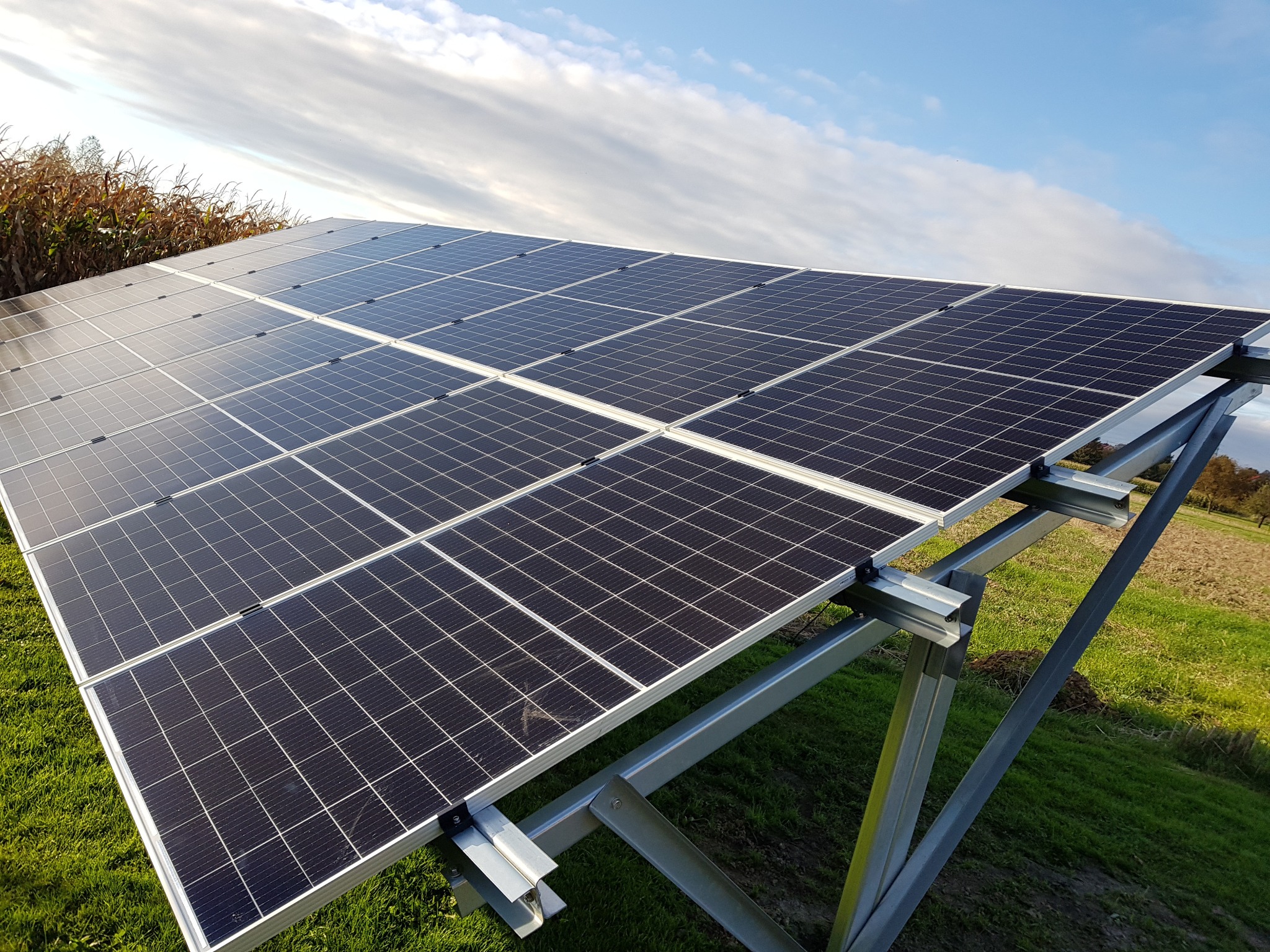 Panel słoneczny na metalowej konstrukcji, widok z dołu pod kątem, na tle zielonej trawy i błękitnego nieba z chmurami.