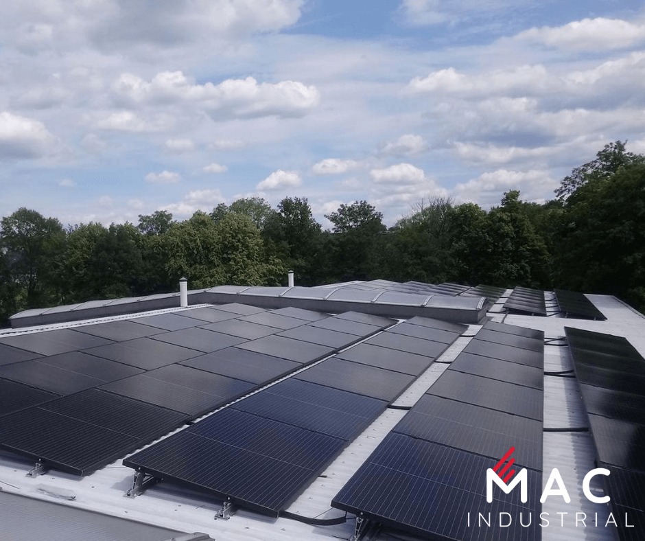 Rząd paneli słonecznych na dachu budynku przemysłowego, widzianych z góry, z logo MAC Industrial w prawym dolnym rogu i drzewami w tle.
