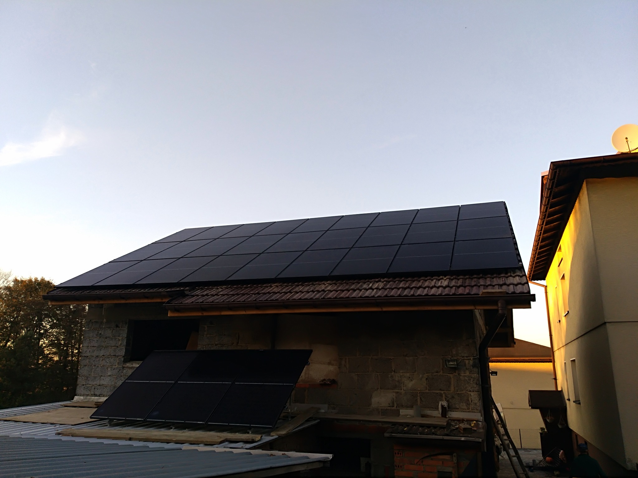 Montaż paneli fotowoltaicznych na dachu surowego budynku z pustaków, widoczna drabina i fragment sąsiedniego budynku z anteną satelitarną w tle.