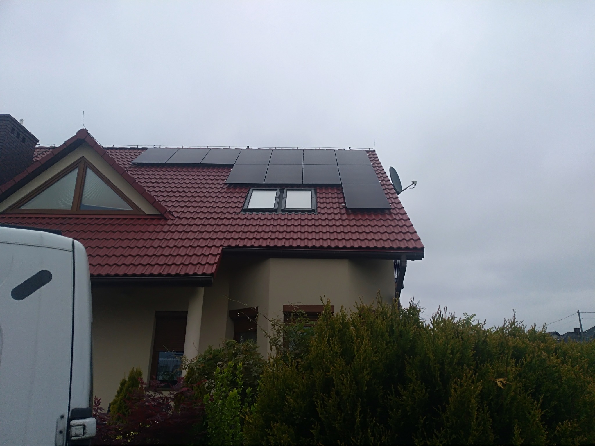 Dom jednorodzinny z czerwoną dachówką i panelami słonecznymi na dachu, widok z poziomu ulicy, częściowo zasłonięty przez krzewy i zaparkowany biały samochód dostawczy.