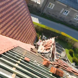 Montaż dachówki karpiówki na drewnianej konstrukcji dachu z widokiem na okoliczną zabudowę i zieleń.